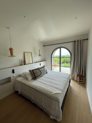 Ein Schlafzimmer mit einem großen Bett, einem Fenster mit Vorhängen, einem kleinen Hocker und einem gerahmten Bild an der Wand.
