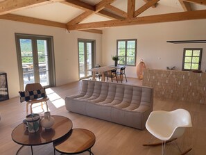 Ein geräumiger Wohnbereich mit einer Couch, einem Esstisch und Stühlen, Holzbalken an der Decke und großen Fenstern.