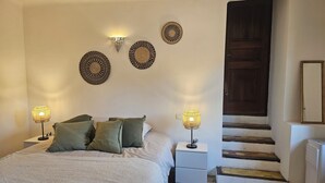 A bedroom with a bed, two bedside lamps, a nightstand, a dresser, a woven rug, and a staircase leading to a dark wooden door. - Small apartment in the village (Lauris)