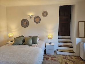 Ein Schlafzimmer mit einem Bett, zwei Nachttischlampen, einem Nachttisch, einer Kommode, einem gewebten Teppich und einer Treppe, die zu einer dunklen Holztür führt.