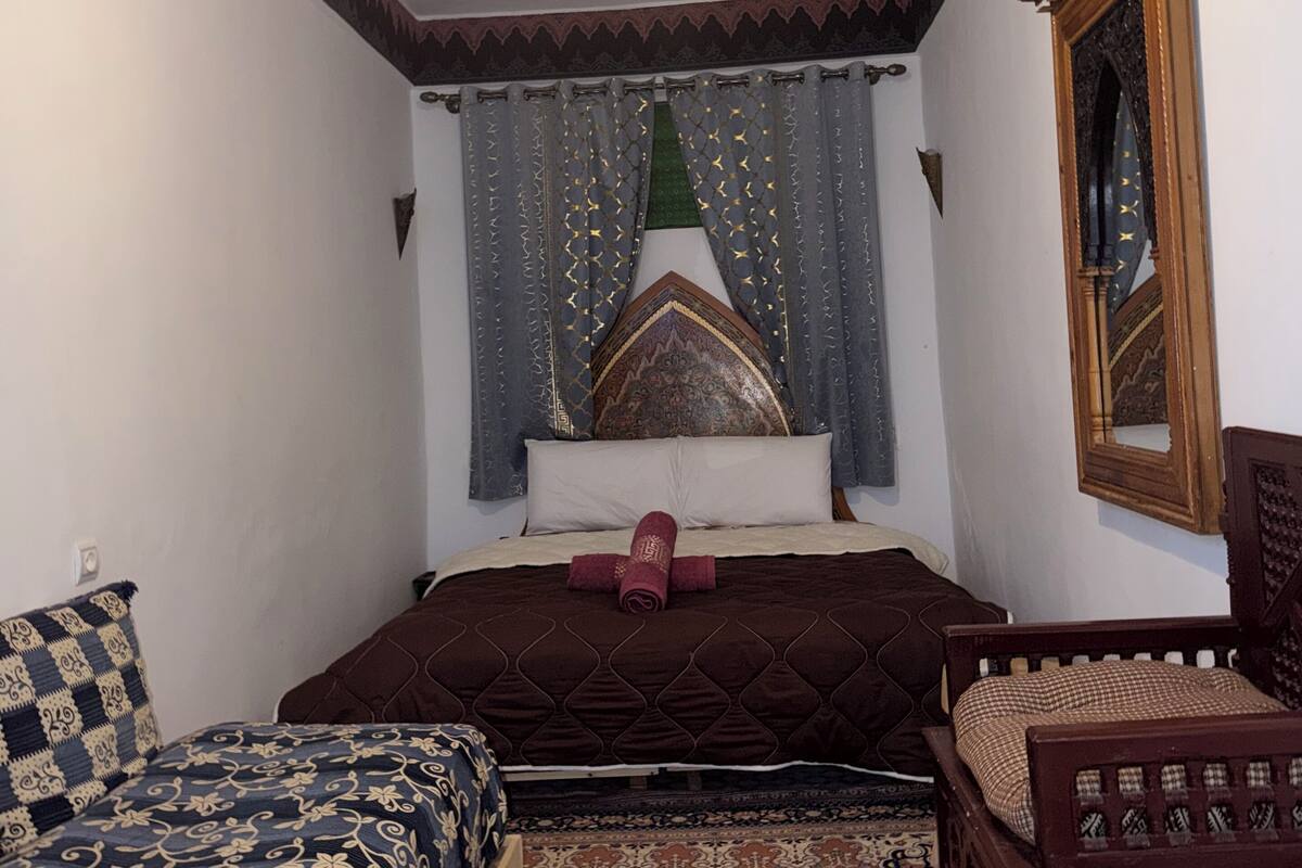 Un dormitorio con una cama, una silla de madera, un tapete estampado, una lámpara decorativa en el techo y un espejo en la pared.