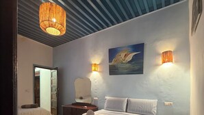 A bedroom with a bed, two bedside tables, a nightstand, a wall-mounted lamp, a painting, and a checkered floor.
