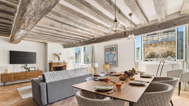 A dining area with a wooden table, cushioned chairs, a sofa, and a television on the wall. - Authentic and Large 1 bdr Flat - Facing Notre Dame (Paris)