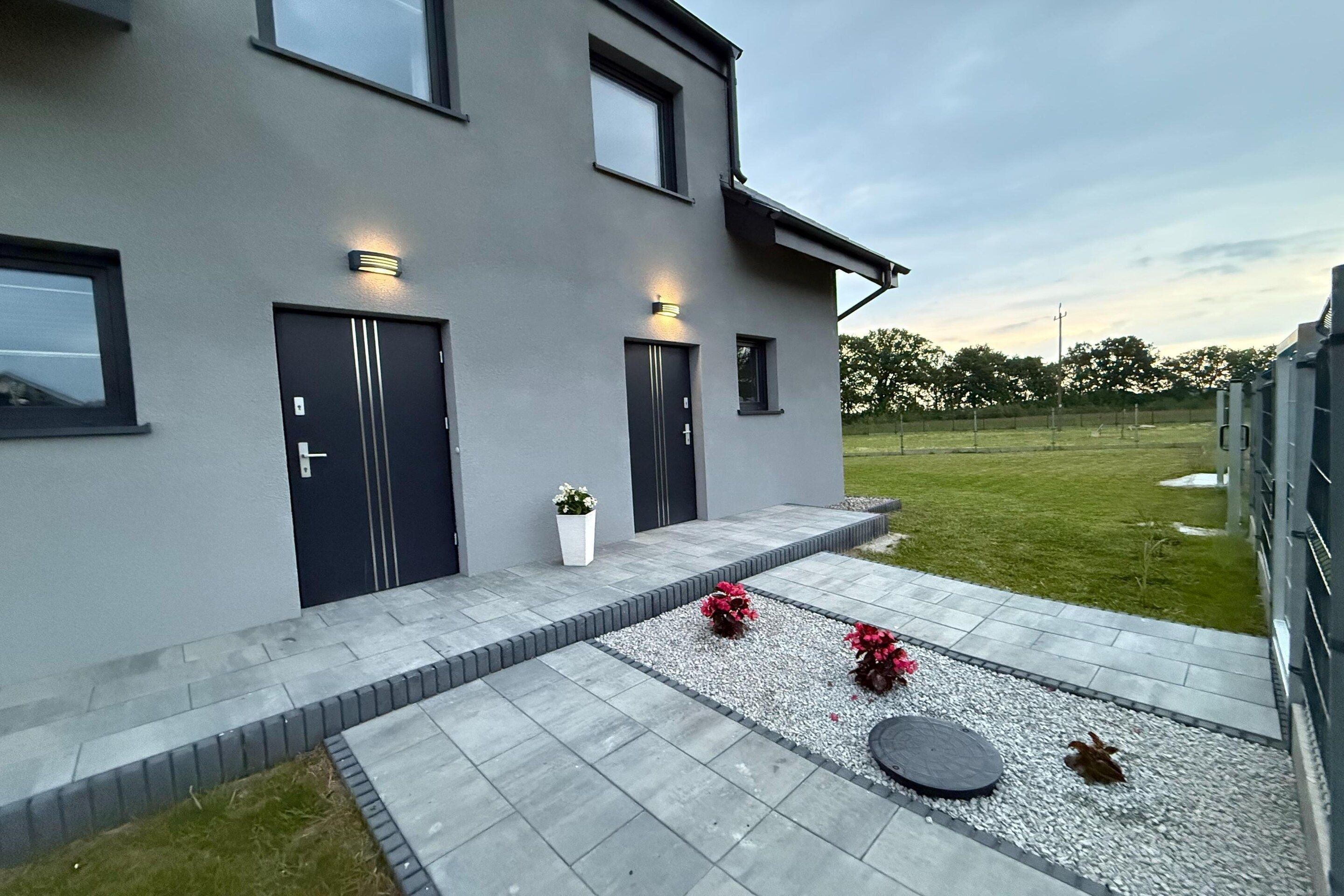 A modern two-story house with a grey exterior, two doors, and a small garden area with gravel and potted plants.