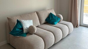 Ein beiges Sofa mit zwei türkisfarbenen Kissen und einem runden Zierkissen, platziert in einem Zimmer mit einem großen Fenster und hellen Fliesen.
