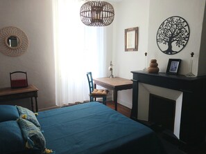 Ein Schlafzimmer mit Bett, Schreibtisch, Stuhl, Kamin und einer Wand mit Spiegel und Kunstwerken.