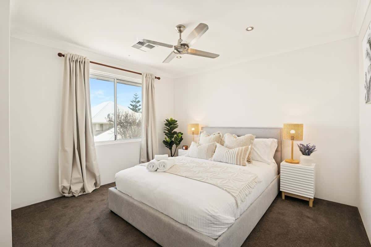 A bedroom with a bed, a ceiling fan, a window with curtains, a bedside table with a lamp, and a plant.