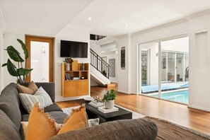 A modern living room with a sofa, coffee table, and bookshelf, a staircase, and a pool view.