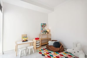 Une salle de jeux pour enfants avec un bureau, des tabourets, un fauteuil poire et un tapis coloré.