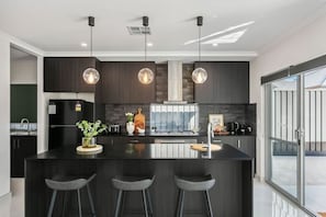 Une cuisine moderne avec des meubles de couleur sombre, un îlot central doté de tabourets de bar, un éclairage suspendu et une vue dégagée grâce à de grandes portes vitrées.