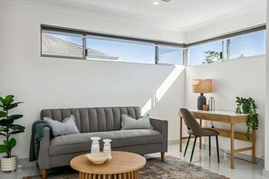 Un salon moderne avec un canapé gris, une table basse ronde en bois, un bureau avec une chaise et une grande fenêtre.
