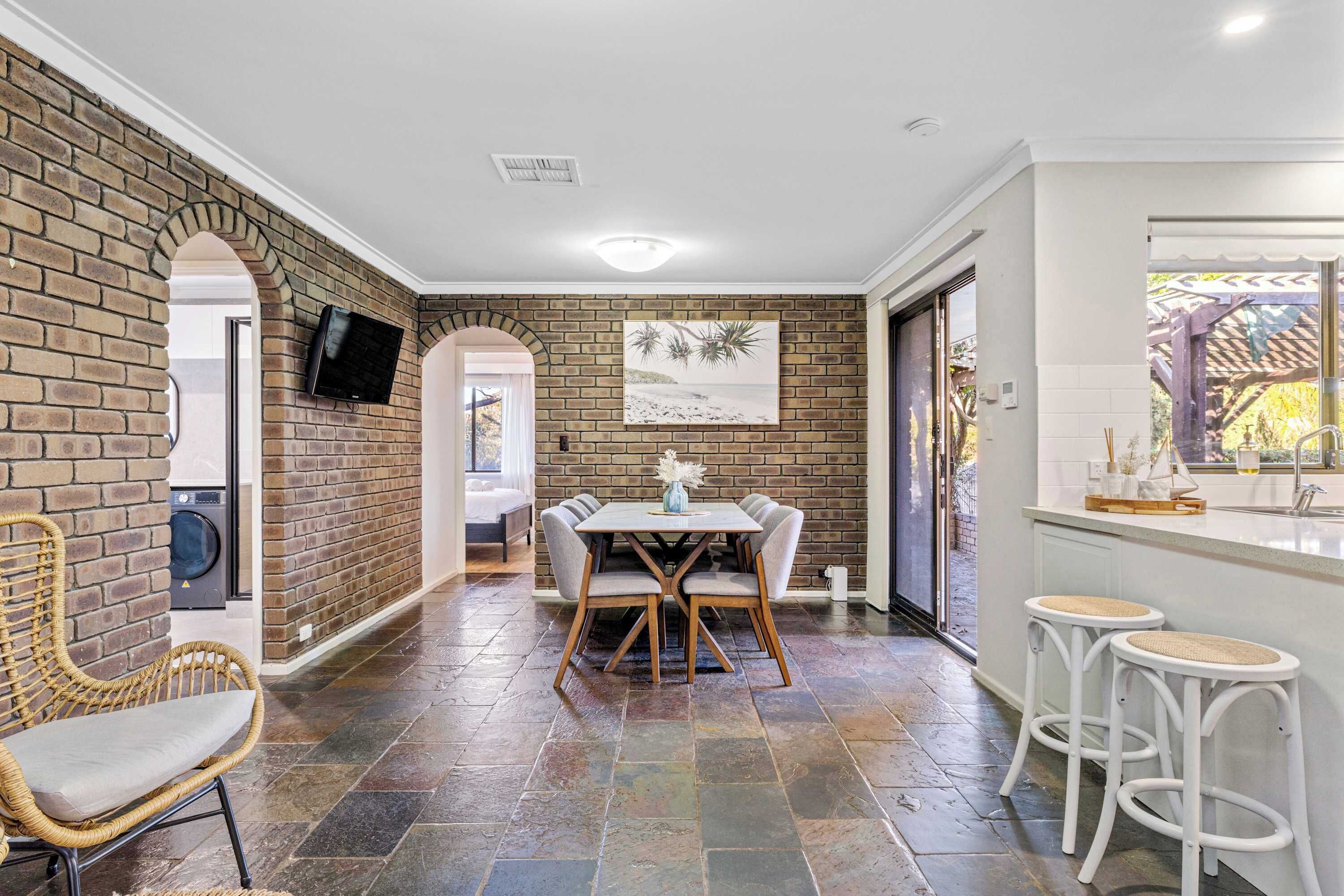 A dining area with a brick wall, a television mounted above a washing machine, and a view of the outdoors through a door.