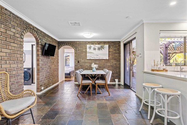 A dining area with a brick wall, a television mounted above a washing machine, and a view of the outdoors through a door.