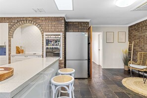 A kitchen with a brick wall, white cabinetry, a built-in oven, and a refrigerator. There are two stools with woven seats and a woven rug on the floor. - Driftwood House | Mullaloo Beach, 3BR + Garden (Mullaloo)