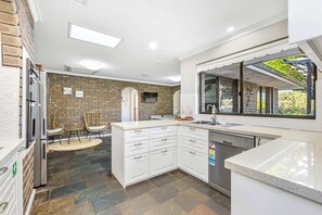A modern kitchen with white cabinetry, a built-in dishwasher, and a large window overlooking a garden. - Driftwood House | Mullaloo Beach, 3BR + Garden (Mullaloo)