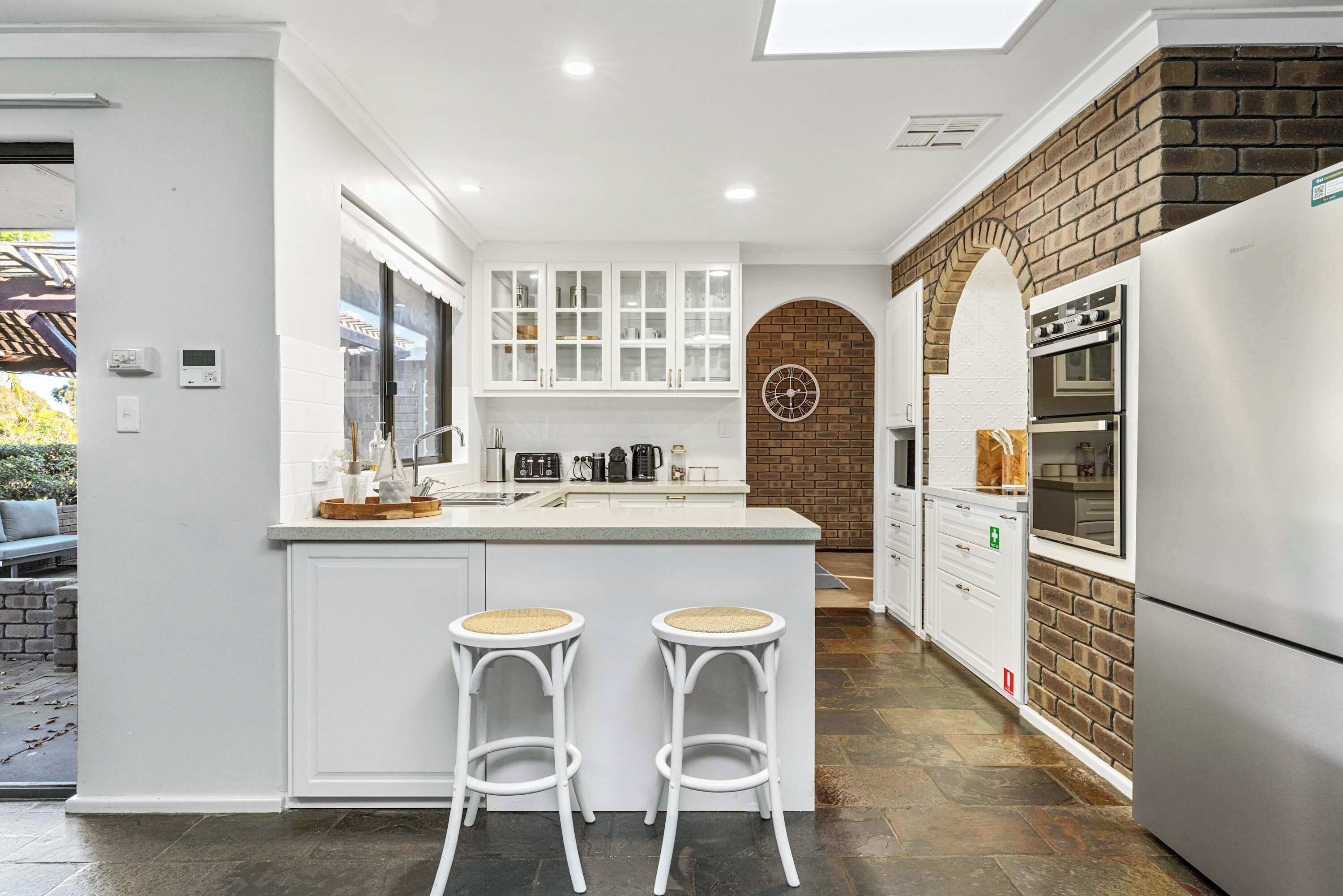 A modern kitchen with white cabinetry, a brick accent wall, a built-in oven, and two bar stools.