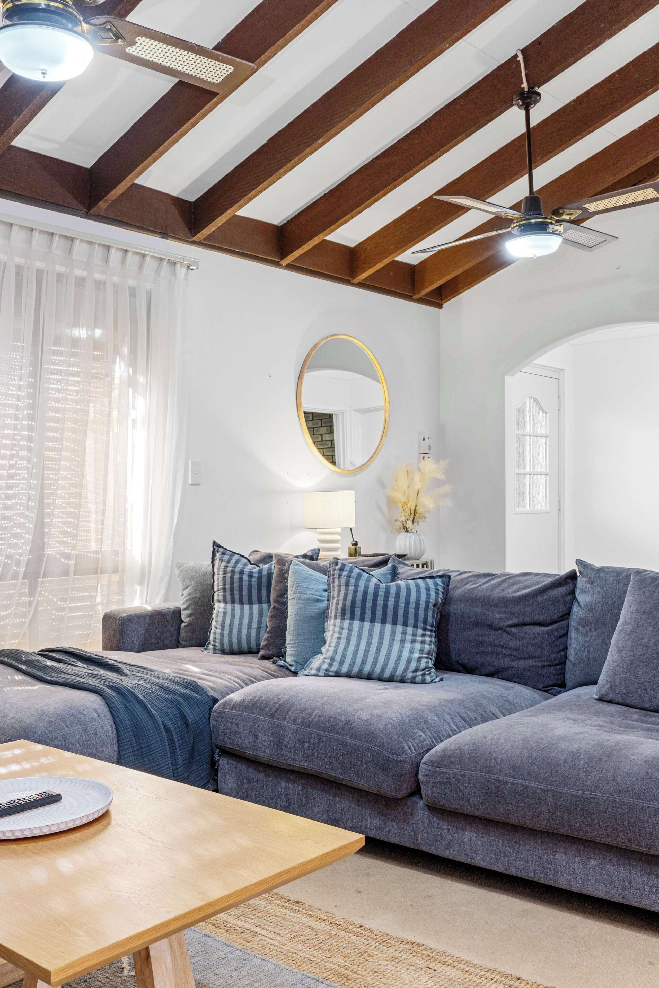 Ein Wohnzimmer mit grauem Sofa, einem Holztisch, einem Deckenventilator und einem großen Fenster mit transparenten Vorhängen.