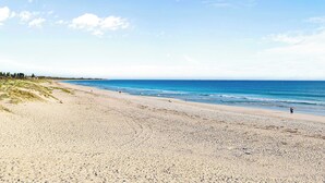 Una playa con cielo despejado, mar tranquilo y algunas personas caminando.