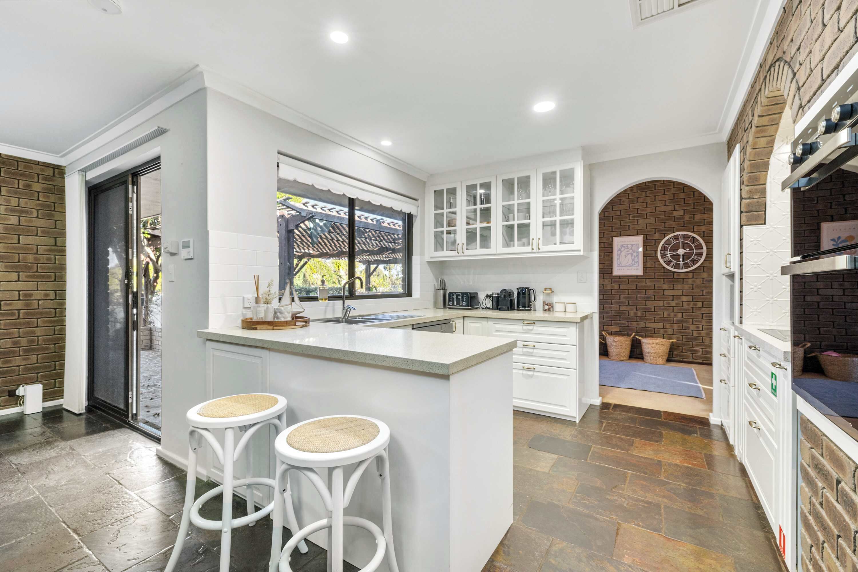 A modern kitchen with white cabinetry, a central island with stools, a brick wall, and a window overlooking greenery.