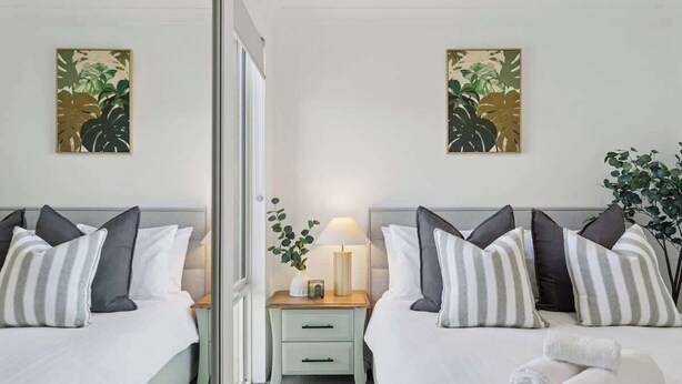 A hotel room with two beds, a nightstand with a lamp, and artwork on the walls. - The Tobias | 4BR, Park-Front + Double Garage (Bayswater)