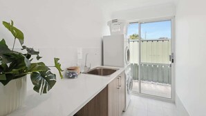 A modern kitchen with a sink, a washing machine, and a sliding glass door leading to an outdoor area. - The Tobias | 4BR, Park-Front + Double Garage (Bayswater)