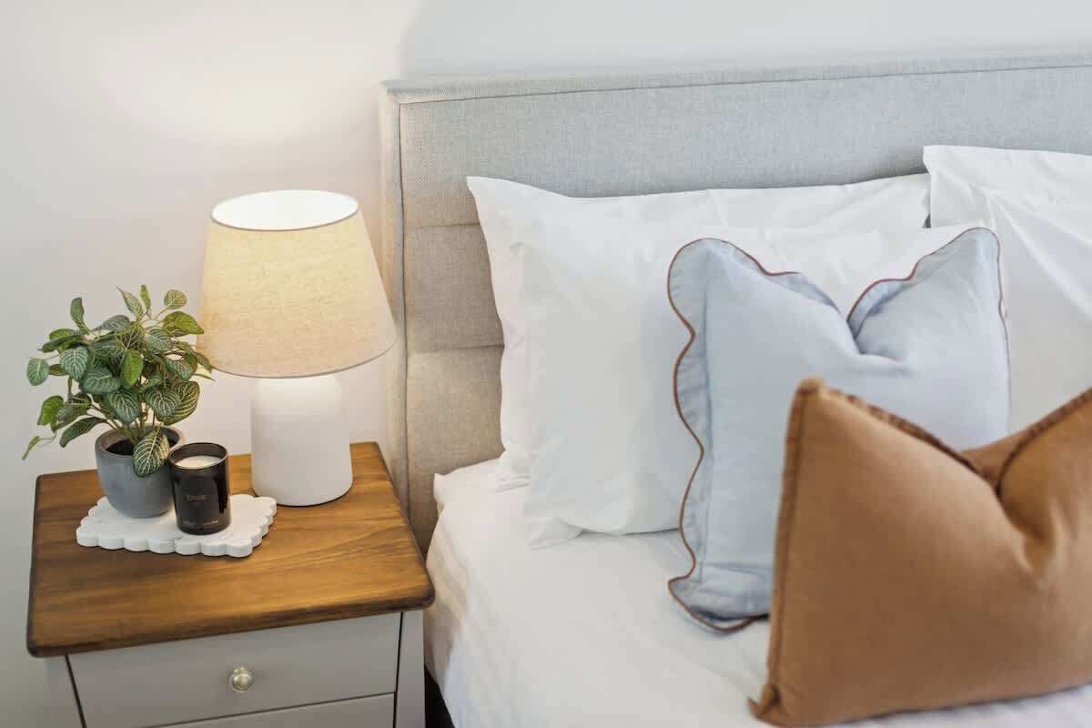 A neatly made bed with white bedding and a few pillows, a bedside table with a lamp, a small potted plant, and a candle.
