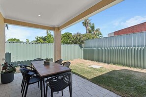 An outdoor patio with a wooden table and chairs, a potted plant, and a green fence.