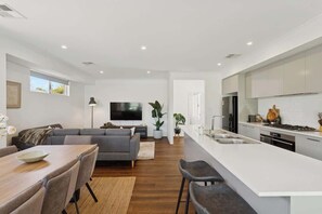 A modern kitchen with a central island, a dining area with a table and chairs, a living room with a sofa and a TV, and a large window providing natural light.