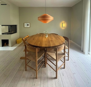 Et rundt spisebord i træ med fire stole, en hængende pendel, en vægmonteret lampe og et indrammet billede på væggen.