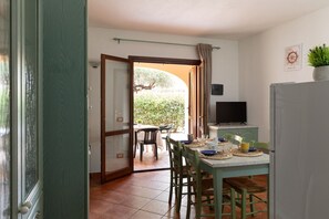 Un comedor con una mesa preparada para cuatro personas, un televisor sobre un soporte y una puerta que da a un espacio exterior.