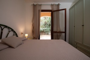 Un dormitorio con cama, mesita de noche con lámpara, armario y puerta que da a un jardín.