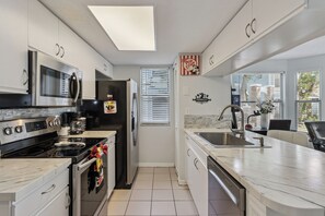Een moderne keuken met roestvrijstalen apparatuur, witte kasten, een groot eiland met een spoelbak en een raam met jaloezieën.