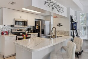 A modern kitchen with stainless steel appliances, a marble countertop island, and a seating area with chairs. - The Magic Hideaway / Disney Escape w/ Pool, Hot Tub & Gym (Kissimmee)