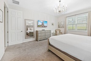 Ein Schlafzimmer mit einem großen Bett, einem Schminktisch, einem Sessel, einer Kristallleuchtre und einem großen Fenster mit Jalousien.