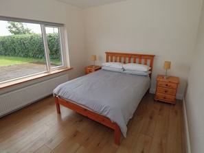 A bedroom with a wooden bed, two bedside tables with lamps, a window with a view of greenery, and a radiator under the window. - Tí Chonaill (Louisburgh)