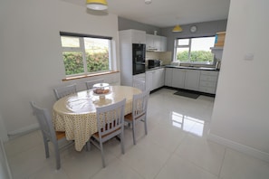 A kitchen with white cabinets, a dining table with chairs, and a window with a view of greenery. - Tí Chonaill (Louisburgh)