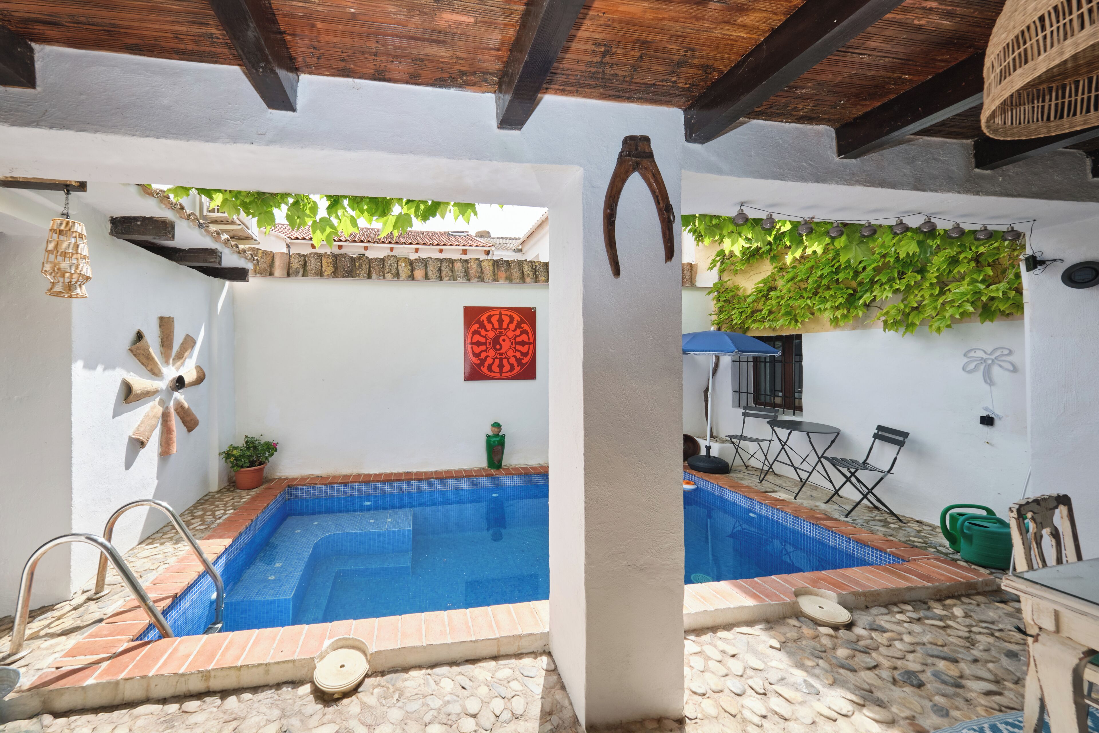 A swimming pool with a tiled edge, a wooden deck, a table with chairs, and a wall with a decorative pattern.. Casa Mathilda