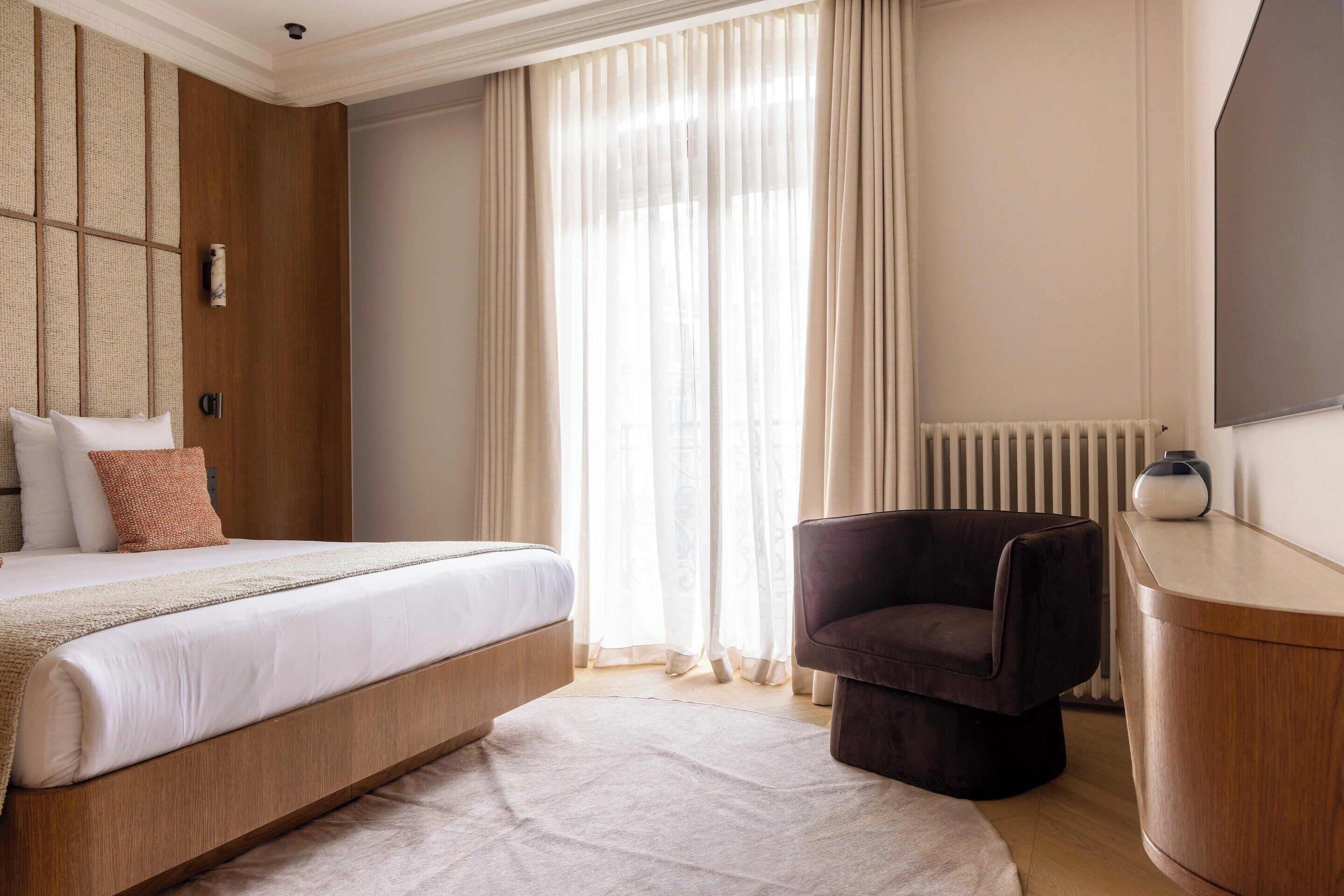 A bedroom with a bed, a chair, a bedside table, and a window with curtains.