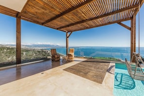 A wooden deck with a view of the sea and mountains, two wooden chairs, and a hammock.