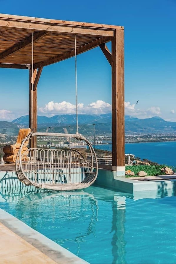 Eine Holzpergola über einem Pool mit einer Hängematte, Blick auf das Meer und Berge im Hintergrund.