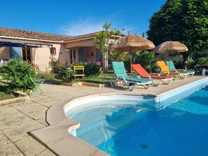 Une piscine avec des chaises longues et des parasols, un bâtiment doté d’un toit en tuiles et un ciel d’un bleu limpide.