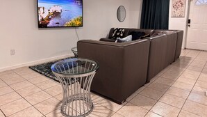 Un saló amb televisió de pantalla plana, una taula de cafè de vidre, un sofà marró, un mirall rodó i una porta amb finestre.