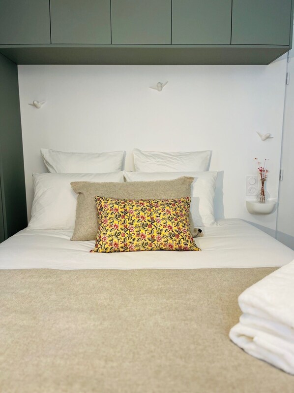 Ein ordentlich bezogenes Bett mit weißen Leinen und einem farbenfrohen, blumigen Kissen, ein kleiner Nachttisch mit einer Vase sowie ein an der Wand befestigtes Regal mit einem dekorativen Gegenstand.