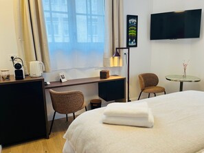 Ein Hotelzimmer mit Bett, Schreibtisch und Stuhl, einem kleinen Tisch mit Vase, einem an der Wand befestigten Fernseher und einem Fenster mit Vorhängen.