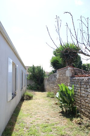 A narrow pathway leading to a building with white walls and shutters, a stone wall with vegetation, and a tree with bare branches. - Les Vagues d'Oléron a 3-bedroom house where you hear the ocean from the garden (Saint-Pierre-d'Oléron)