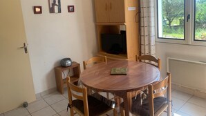 Un comedor con una mesa redonda de madera, seis sillas, un televisor sobre un mueble de madera y un ventanal con vista a árboles.
