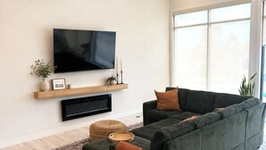 A modern living room with a sectional sofa, a flat-screen TV, a wooden shelf, and a large window.