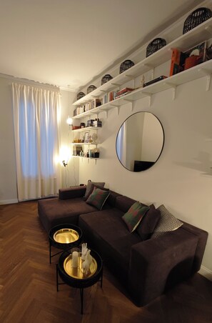 A living room with a sectional sofa, two side tables, a round mirror, and shelves with books and decorative items. - BB Suite Florence – Elegance, Comfort, Hospitality. (Firenze)