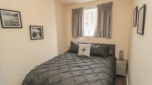 A bedroom with a bed, a window with curtains, a bedside table with a lamp, and two framed pictures on the wall. - Memory Maker (Bridlington)