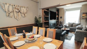 A dining area with a wooden table set for six, surrounded by chairs. There is a television in the background and a seating area with sofas. - Memory Maker (Bridlington)